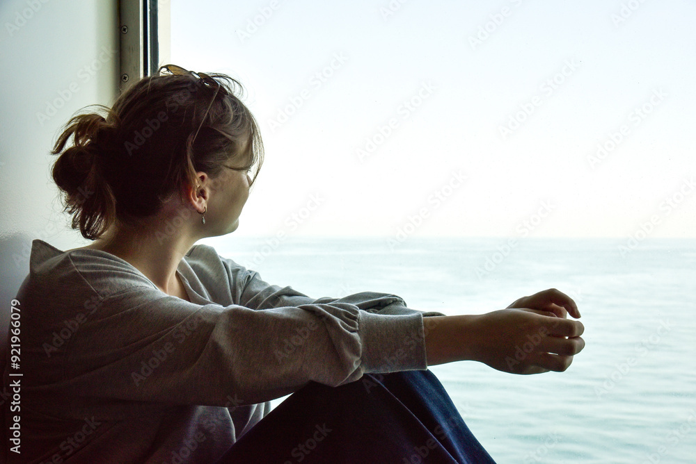 young woman in a grey shirt and jeans is sitting by a window looking at the sea