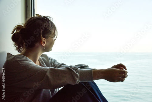Obraz na płótnie young woman in a grey shirt and jeans is sitting by a window looking at the sea w salonie