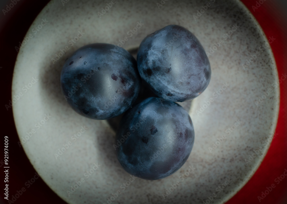 A close-up photograph of three ripe, dark blue plums arranged on a light ceramic plate with a subtle texture. A close-up photograph of three ripe, dark blue plums arranged on a light ceramic plate with a subtle texture.