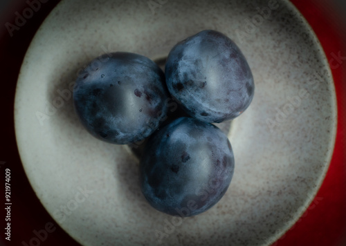 A close-up photograph of three ripe, dark blue plums arranged on a light ceramic plate with a subtle texture. 