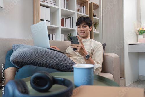 Young Man Relaxing in Modern Room with Laptop and Smartphone, Enjoying Leisure Time on Comfortable Couch