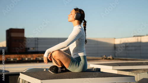 Young sporty woman with big headphones practicing lotus asana. Woman do meditation yoga practice with eyes closed alone. Well being, wellness concept