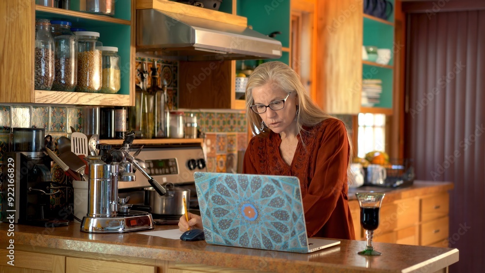 Mature woman writing notes on paper while looking at computer screen in kitchen. Mature woman writing notes on paper while looking at computer screen in kitchen.