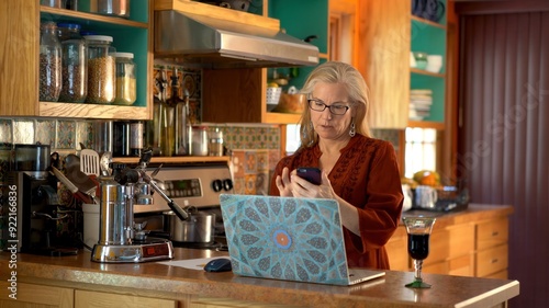 Dialing phone, a mature woman types on computer setting on a kitchen counter.