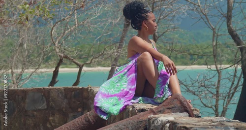 A young lady, dressed in a long, flowing gown, stands on a cliff with the beach spread out beneath her. photo