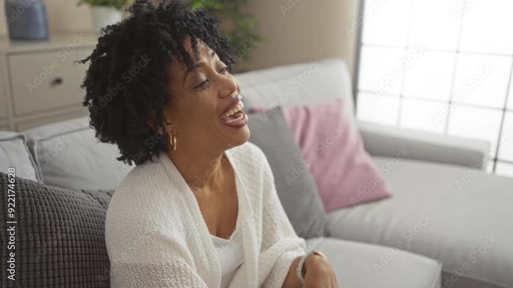 A beautiful young african american woman with curly hair is smiling and laughing in a comfortable living room setting with cozy cushions on a couch. A beautiful young african american woman with curly hair is smiling and laughing in a comfortable living room setting with cozy cushions on a couch.