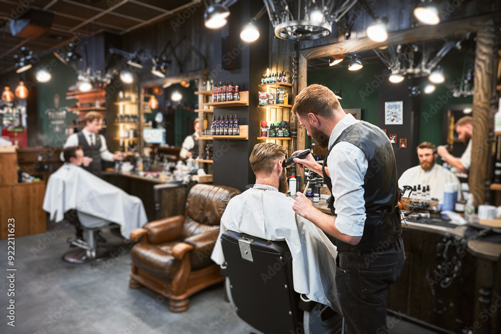 Professional barber using comb and electric shaver to cut side hair. Male customer getting stylish haircut. Side view of man on background of other hairdresser. Professional barber using comb and electric shaver to cut side hair. Male customer getting stylish haircut. Side view of man on background of other hairdresser.