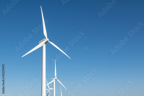 A row of tall wind turbines stand against a clear blue sky, with the sun shining brightly and a small moon visible.