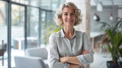 Confident businesswoman with crossed arms smiling in modern office, facing camera, embodying success in professional attire with sleek hairstyle.