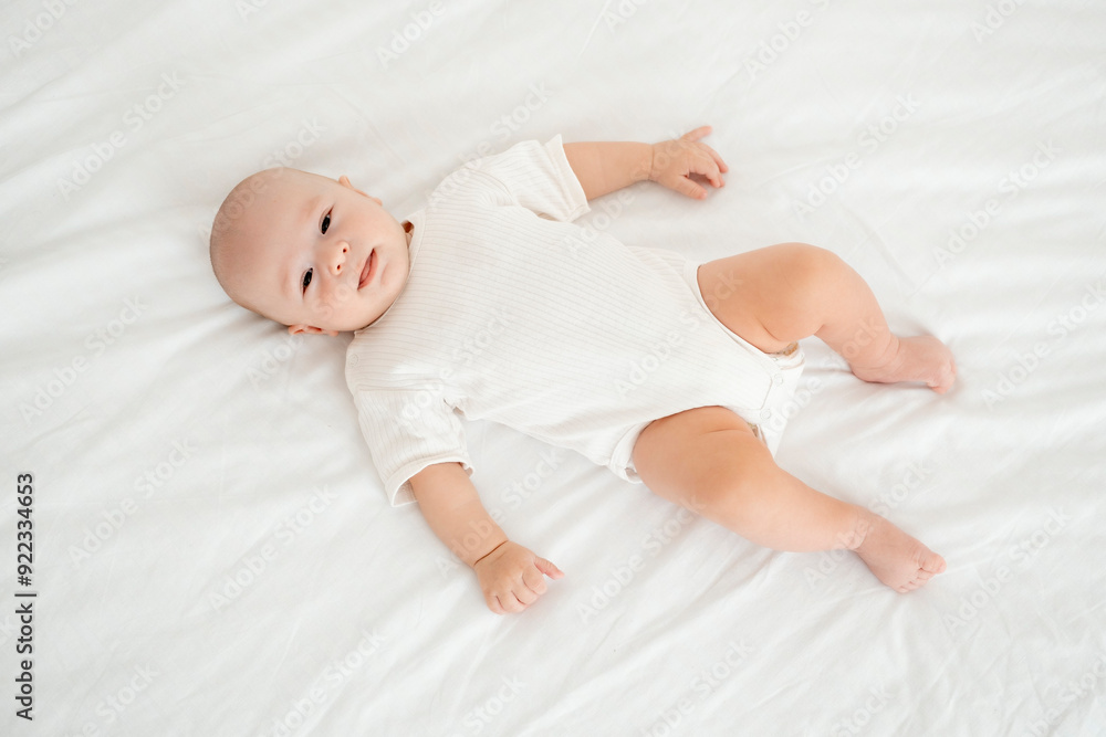 a baby in a white bodysuit on the bed at home smiling, a portrait of a baby on the bed, a small child going to bed, a place for text a baby in a white bodysuit on the bed at home smiling, a portrait of a baby on the bed, a small child going to bed, a place for text