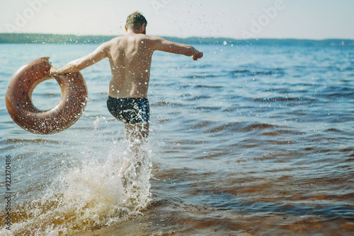 man having fun running into lake holding a rubberswimming ring behaving like a child. regress to childhood, grow childish concept photo