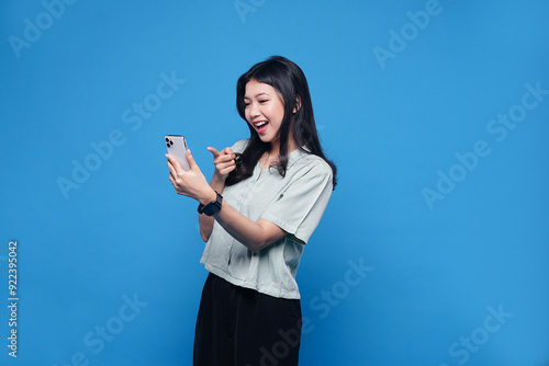 Happy Asian young woman using mobile smartphone, read good news online, laugh excited getting offer, great positive surprise, celebrate success on smartphone. Smile girl isolated on blue background
