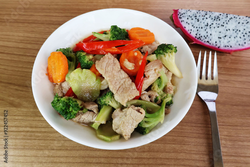 Stir-fried pork, broccoli, carrots, chili in a porcelain plate. Place a fork on the side. and sliced ​​dragon fruit on a wooden table