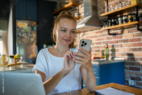 Young woman using smartphone in kitchen while working from home