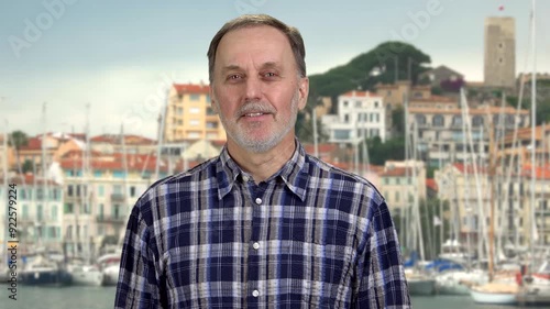 Smiling handsome casual-dressed caucasian aged senior man standing outdoors. European town with yacht harbour in the background. photo