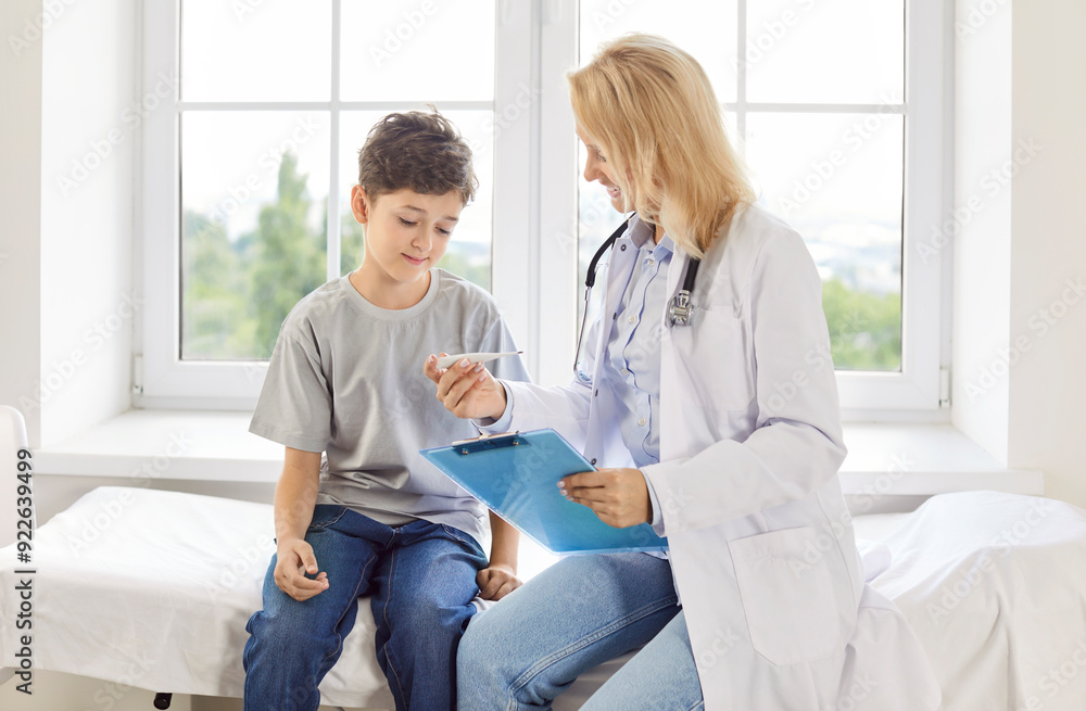 Little sick boy patient sitting on couch in medical clinic with friendly young female pediatrician doctor and looking on thermometer during medical exam. Child health care and medicine concept. Little sick boy patient sitting on couch in medical clinic with friendly young female pediatrician doctor and looking on thermometer during medical exam. Child health care and medicine concept.