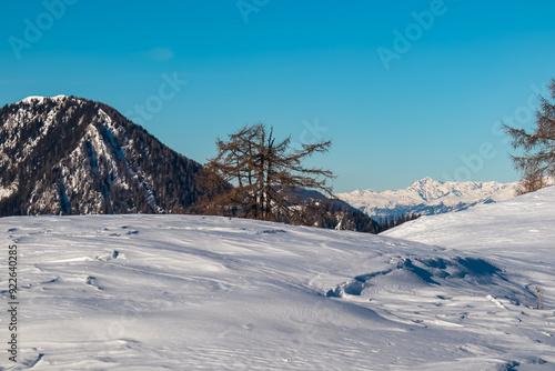 Winter landscape in Bleiberger Eisenerz, Carinthia, Austrian Alps. Snow-covered terrain stretches towards majestic mountain range Julian Alps in warm glow of midday sun. Footprints and ski tracks photo