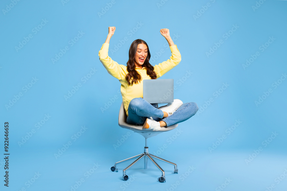 Excited European lady sitting on chair with laptop and celebrating success against blue background. Woman enjoying big sale or online win Excited European lady sitting on chair with laptop and celebrating success against blue background. Woman enjoying big sale or online win