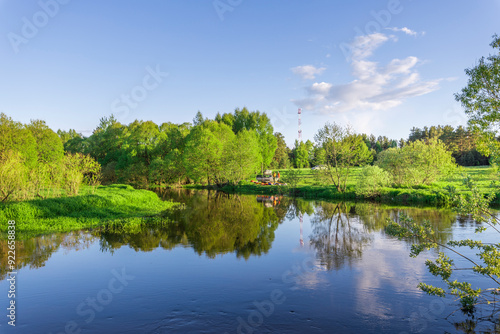 A calm river reflects the vibrant green foliage under a clear blue sky on a beautiful spring afternoon, inviting moments of peace and tranquility.