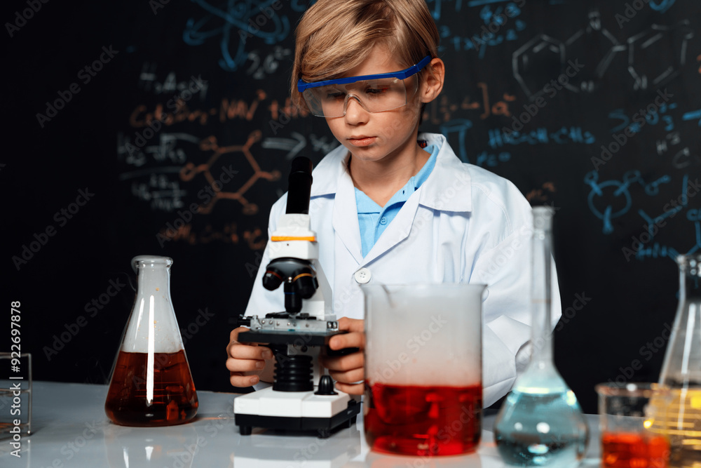 Blonde hair schoolboy in laboratory wear lab coat stand and learn science of chemistry technology in STEM class. The student looking object in microscope . On the table put many flask. Erudition. Blonde hair schoolboy in laboratory wear lab coat stand and learn science of chemistry technology in STEM class. The student looking object in microscope . On the table put many flask. Erudition.