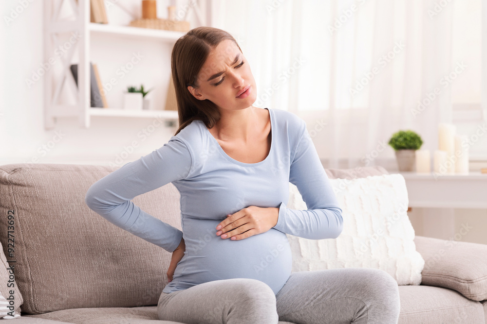 Backache. Pregnant Girl Suffering From Pain In Lower Back And Spasms Sitting On Couch At Home. Backache. Pregnant Girl Suffering From Pain In Lower Back And Spasms Sitting On Couch At Home.