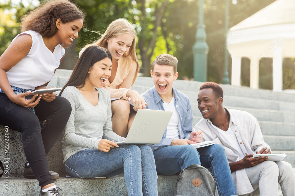 Excited multiracial friends checking test results on laptop online, sitting on stairs in public park, empty space Excited multiracial friends checking test results on laptop online, sitting on stairs in public park, empty space