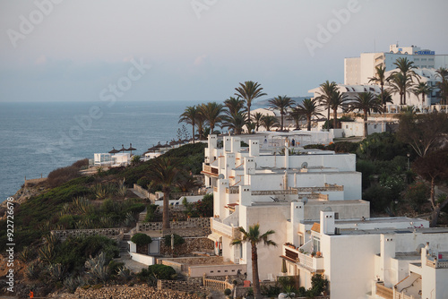 A serene village of whitewashed buildings with flat roofs, nestled among palm trees. The calm sea and soft lighting enhance the Mediterranean atmosphere.