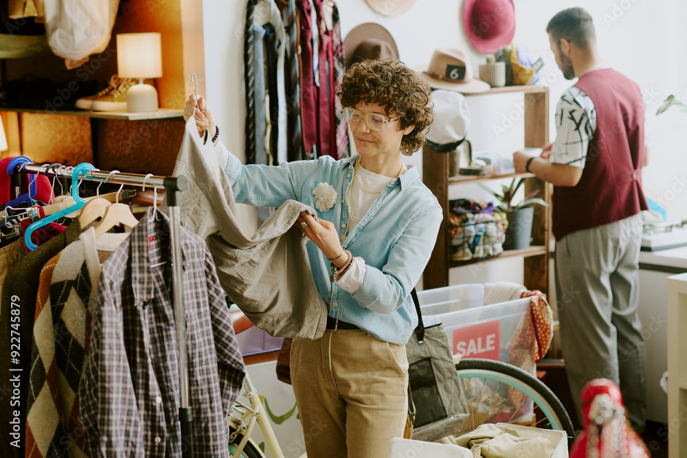 © AnnaStills - Man with curly hair examining clothing item in vibrant vintage store while another person browses items in background. Environments filled with diverse articles and sale signs