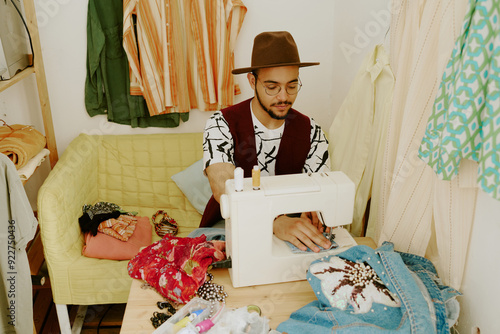 Φωτογραφία Man wearing hat operating sewing machine in cozy room filled with various garmen