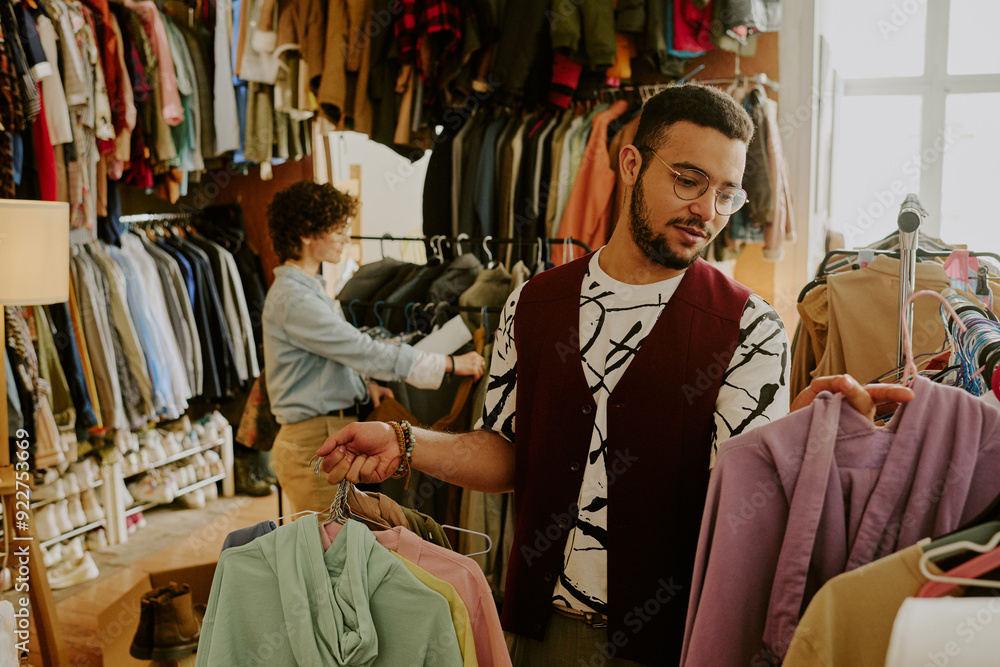 Man carefully selecting clothing in modern boutique while putting garment on rack with concentration, shelves with folded clothes and woman browsing in background Man carefully selecting clothing in modern boutique while putting garment on rack with concentration, shelves with folded clothes and woman browsing in background