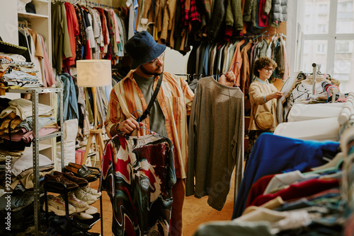 Person browsing racks of clothing in bright thrift store with various garments on display during daylight Peering at different shirts and examining styles and options