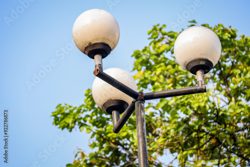 A close-up of a modern street lamp with three round bulbs, set against a clear blue sky and green foliage. The slightly rusted metal adds a touch of urban charm photo