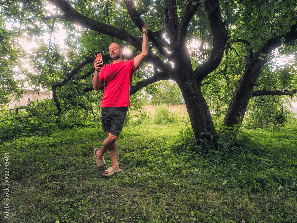A man in a red shirt stands in shade in front of a tree. The image has a peaceful and serene mood, as the man is standing in a lush green garden on a hot summer day. Country life and holiday theme. A man in a red shirt stands in shade in front of a tree. The image has a peaceful and serene mood, as the man is standing in a lush green garden on a hot summer day. Country life and holiday theme.