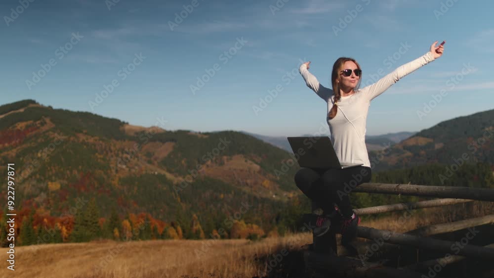 Young woman is sitting on a wooden fence with a laptop on her lap and raising her hands in the air enjoying the mountain scenery