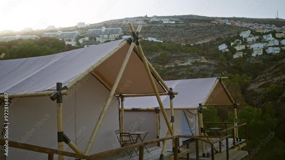 Bamboo-constructed tents set up on wooden platforms in a serene hillside landscape with white houses and lush greenery in the background during a camping adventure. Bamboo-constructed tents set up on wooden platforms in a serene hillside landscape with white houses and lush greenery in the background during a camping adventure.