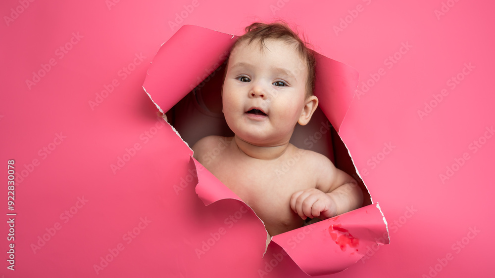 Cute Caucasian newborn baby boy peeks out of a hole in a paper pink background. Cute Caucasian newborn baby boy peeks out of a hole in a paper pink background.