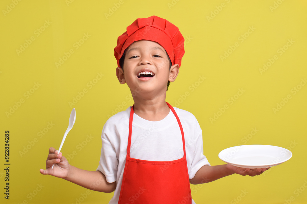 Cute little chef with kitchen plate and spoon. Asian boy wearing red apron and hat on isolated yellow background Cute little chef with kitchen plate and spoon. Asian boy wearing red apron and hat on isolated yellow background