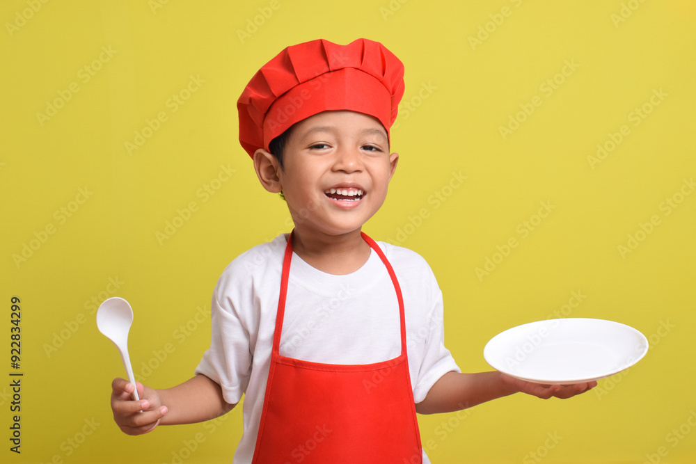 Cute little chef with kitchen plate and spoon. Asian boy wearing red apron and hat on isolated yellow background Cute little chef with kitchen plate and spoon. Asian boy wearing red apron and hat on isolated yellow background