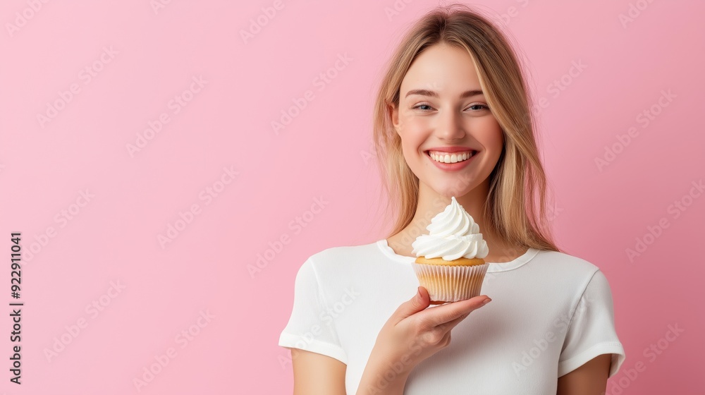 Young woman smiles while holding a frosted cupcake against a pink backdrop. Young woman smiles while holding a frosted cupcake against a pink backdrop.