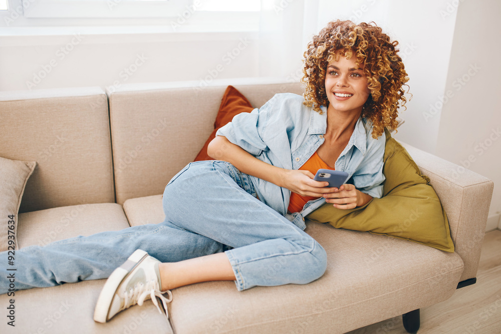Obraz premium Happy woman holding mobile phone and relaxing on cozy sofa at home