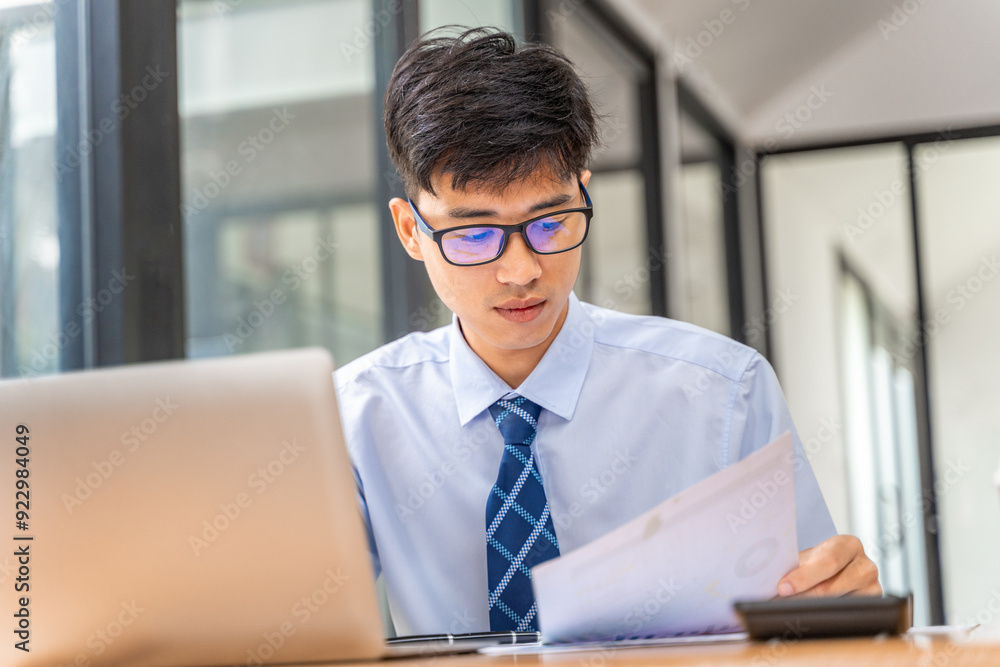 Young businessman working on his project in his office checking business result data from the printed sheets and his laptop computer Young businessman working on his project in his office checking business result data from the printed sheets and his laptop computer