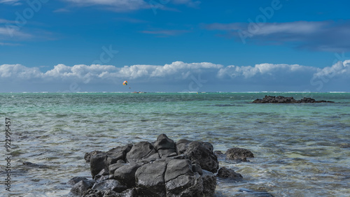 The endless turquoise ocean. A multicolored parachute, tiny silhouettes of flying people are visible in the blue sky. Boats at sea. In the foreground is a pile of picturesque black volcanic boulders photo