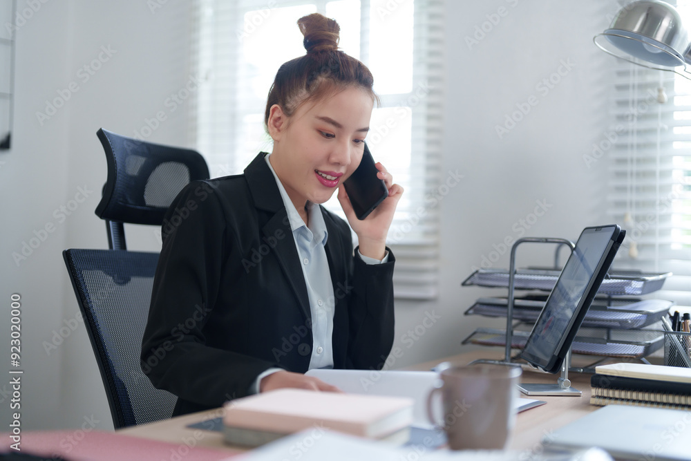 A businesswoman in a black suit is talking on the phone while working at her desk with a laptop, embodying multitasking and efficiency in a professional office setting A businesswoman in a black suit is talking on the phone while working at her desk with a laptop, embodying multitasking and efficiency in a professional office setting