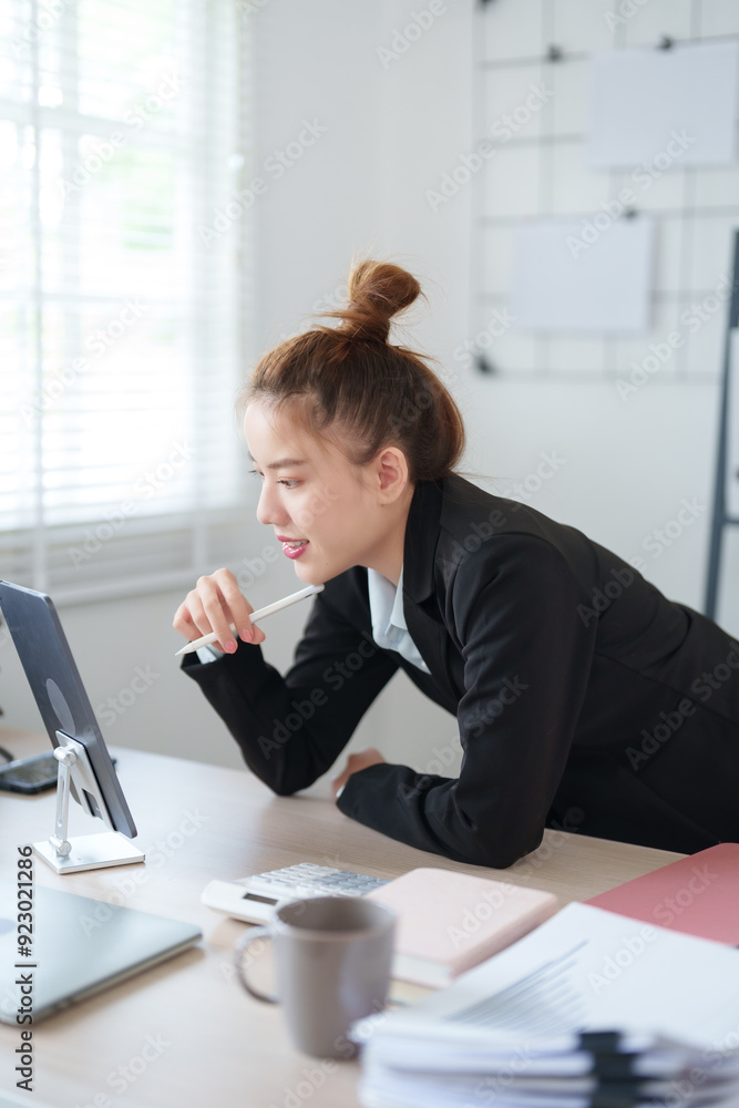 A businesswoman in a black suit is leaning over her desk, deeply focused on her work in a modern office environment
