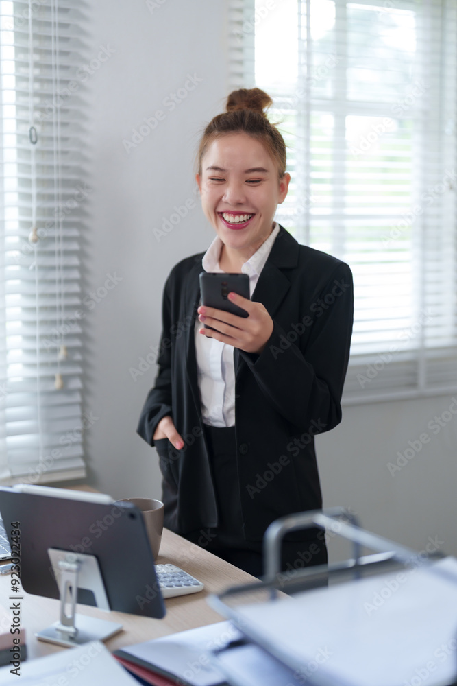A businesswoman in a black suit is standing by the window, checking her smartphone with a focused expression in a modern office setting A businesswoman in a black suit is standing by the window, checking her smartphone with a focused expression in a modern office setting