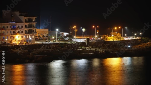 Night timelapse from Zonqor beach with illuminated buildings and a parking lot, and with people on the seafront in Marsaskala, Malta. photo