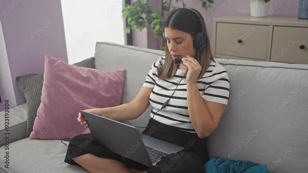 Young hispanic woman wearing headphones works from home on a laptop while sitting on a cozy grey sofa in a stylish living room with pink and grey cushions, relaxed and focused on her task. Young hispanic woman wearing headphones works from home on a laptop while sitting on a cozy grey sofa in a stylish living room with pink and grey cushions, relaxed and focused on her task.