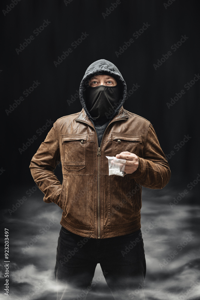Drug dealer, dangerous hooded criminal man in black mask and brown leather jacket holding transparent plastic bag with drugs, on a dark background with smoke Drug dealer, dangerous hooded criminal man in black mask and brown leather jacket holding transparent plastic bag with drugs, on a dark background with smoke