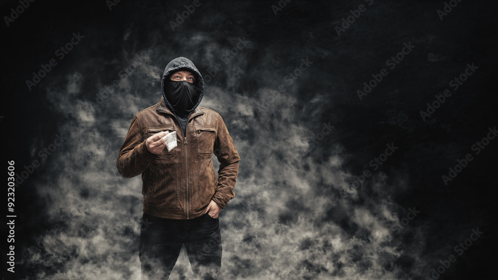 Drug dealer, dangerous hooded criminal man in black mask and brown leather jacket holding transparent plastic bag with drugs, on a dark background with smoke Drug dealer, dangerous hooded criminal man in black mask and brown leather jacket holding transparent plastic bag with drugs, on a dark background with smoke