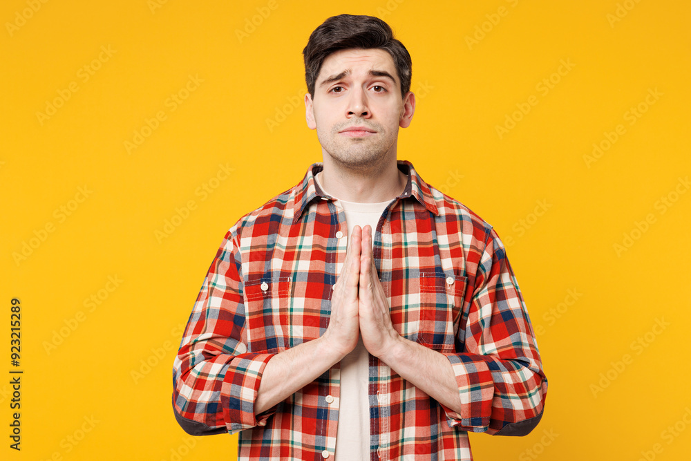 Young Caucasian man he wears red checkered shirt casual clothes hold hands folded in prayer gesture, begging about something, look camera isolated on plain yellow orange background. Lifestyle concept. Young Caucasian man he wears red checkered shirt casual clothes hold hands folded in prayer gesture, begging about something, look camera isolated on plain yellow orange background. Lifestyle concept.